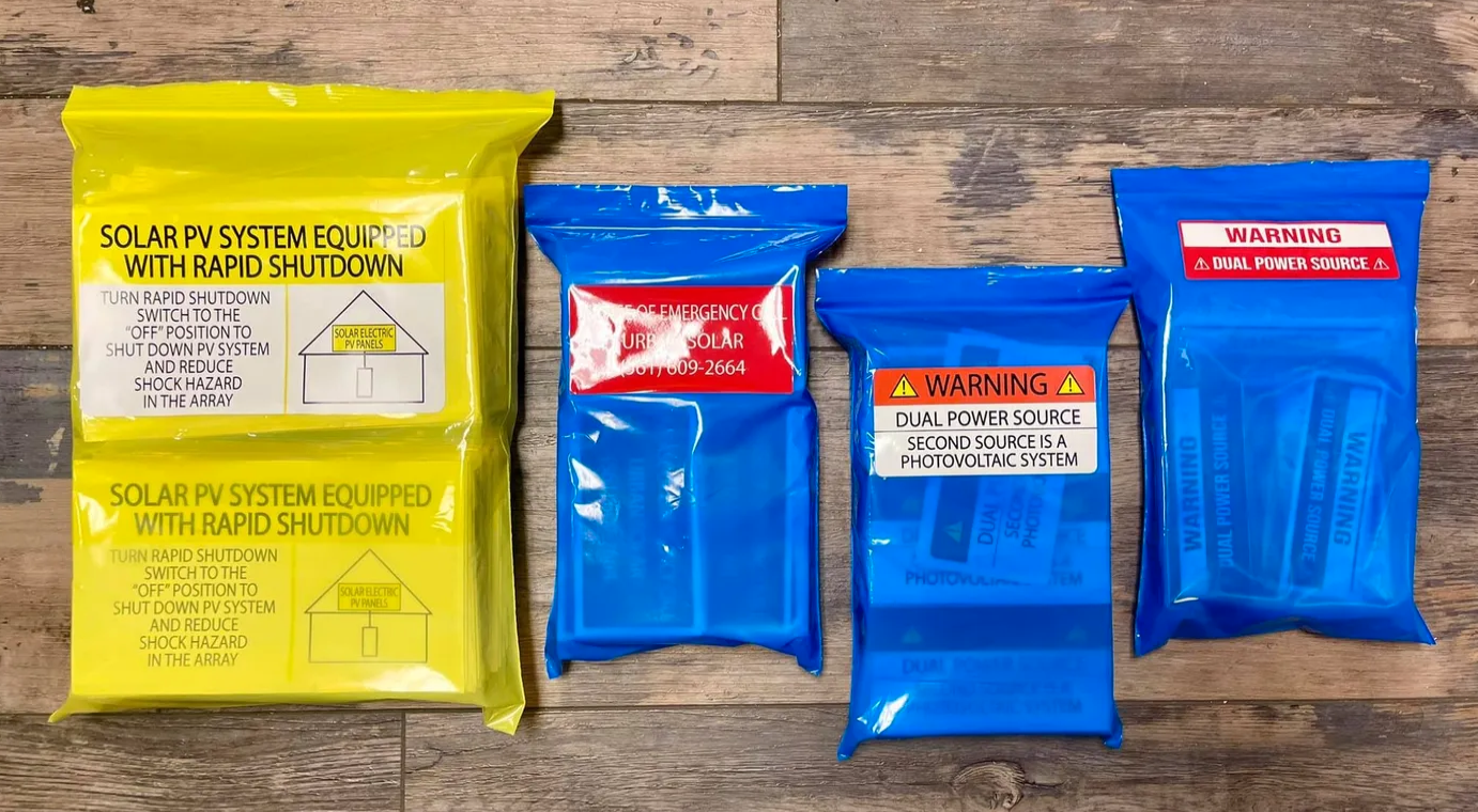 Assorted solar photovoltaic safety labels in yellow and blue plastic bags, including large yellow Rapid Shutdown and red emergency contact labels, custom-printed by BAPS Signs & Wraps in Oldsmar, FL