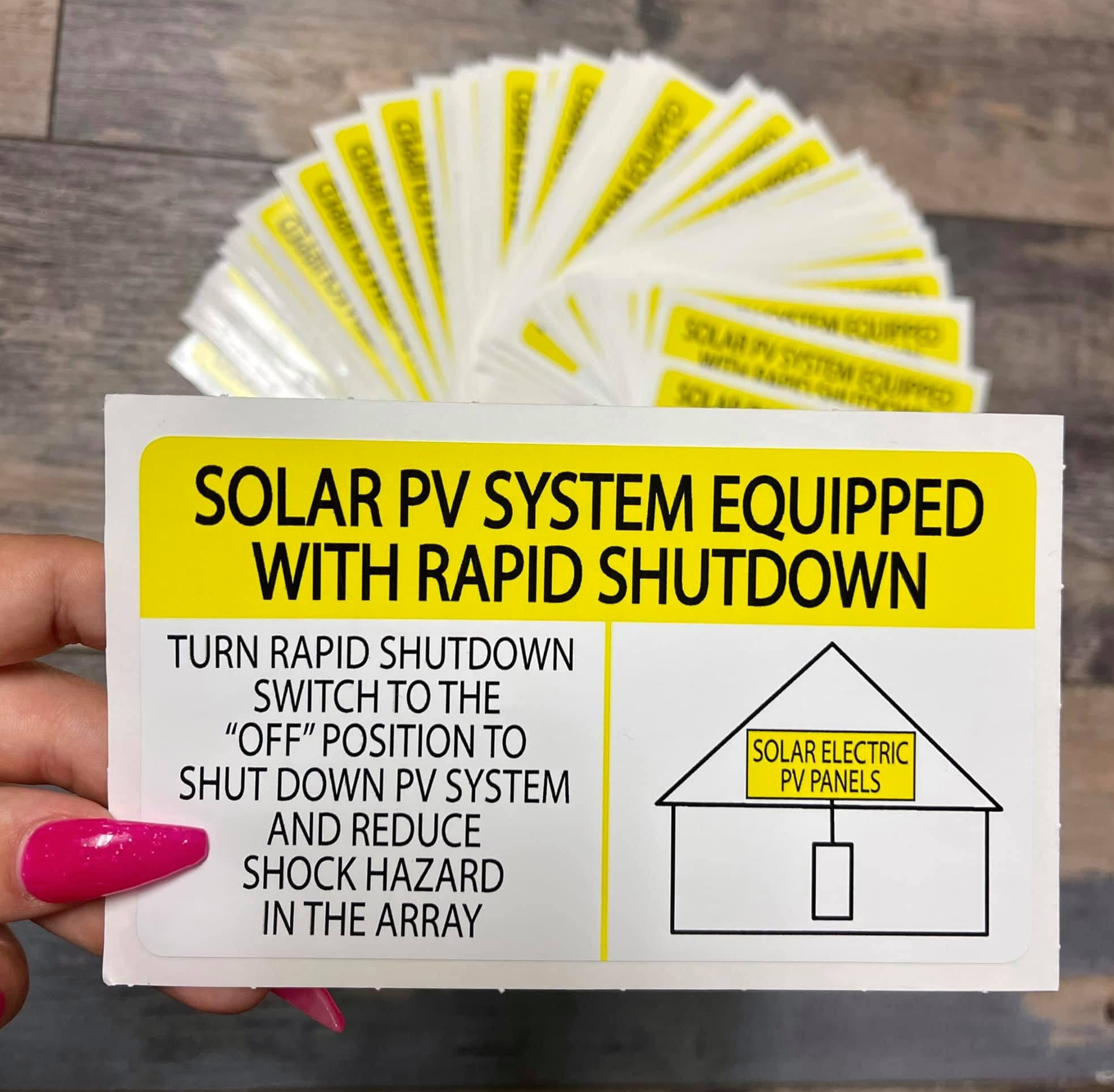 Hand holding a white and yellow label reading SOLAR PV SYSTEM EQUIPPED WITH RAPID SHUTDOWN with instructions and a house diagram, custom-printed by BAPS Signs & Wraps in Oldsmar, FL
