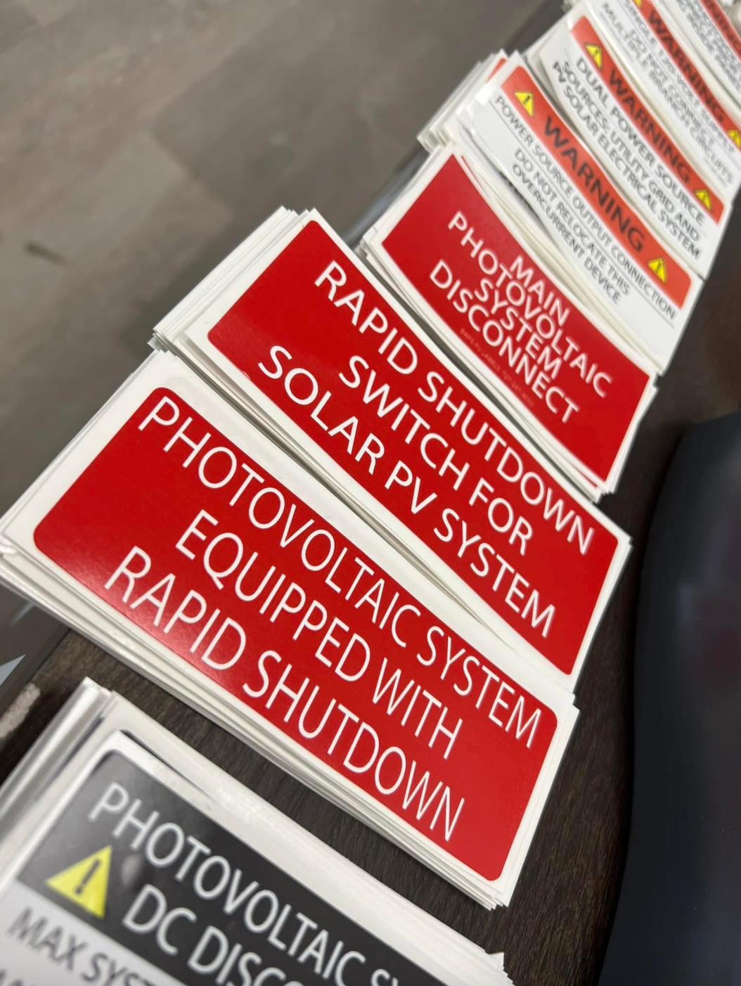 Stacks of red, white, and black adhesive solar safety labels including Rapid Shutdown Switch and Main Photovoltaic System Disconnect labels, custom-printed by BAPS Signs & Wraps in Oldsmar, FL