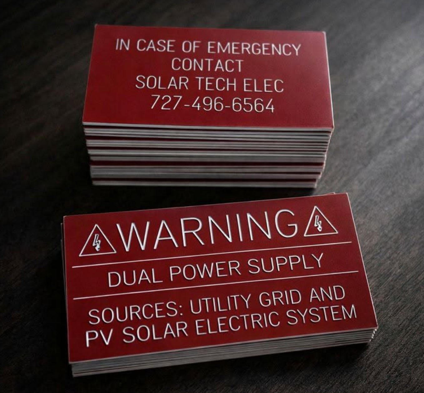 Two stacks of red engraved-style plaques for Solar Tech Elec — an emergency contact plaque with phone number 727-496-6564 and a dual power supply warning plaque, custom-printed by BAPS Signs & Wraps in Oldsmar, FL